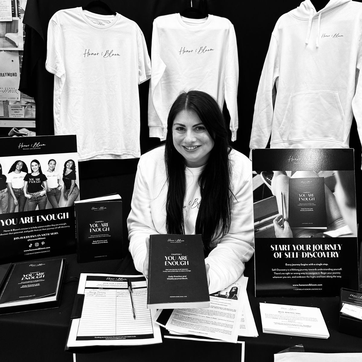 Woman sitting at a table with branded merchandise including t-shirts, hoodies, and brochures.