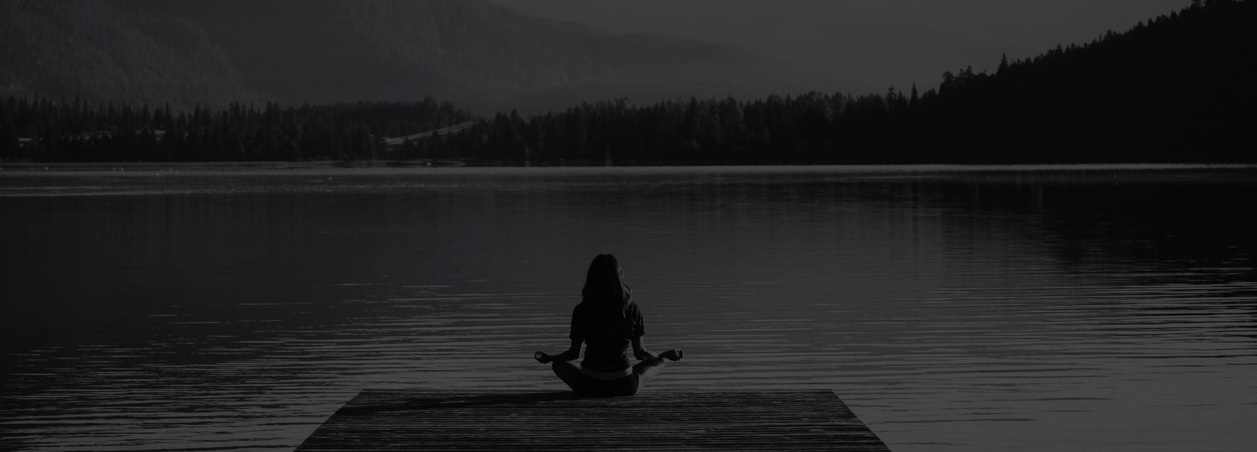 Person meditating on a dock by a lake with mountains in the background.